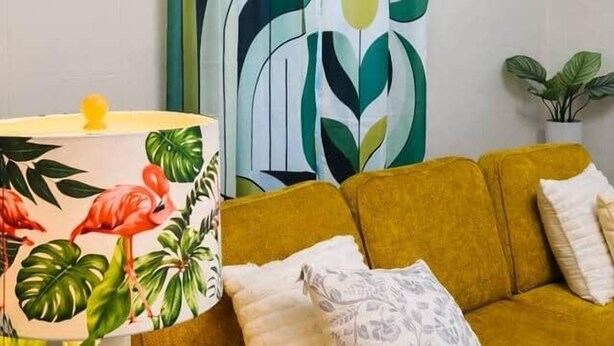 Une pièce avec un canapé jaune, une lampe à motifs, un rideau abstrait vert et blanc, et des étagères murales ornées de plantes.