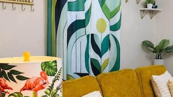 Una habitación con un sofá amarillo, una lámpara estampada, una cortina abstracta verde y blanca, y estanterías con plantas.