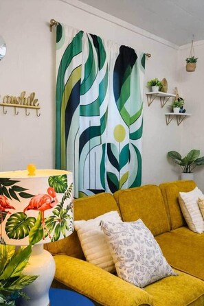 A room with a yellow sofa, a patterned lamp, a green and white abstract curtain, and wall-mounted shelves with plants.