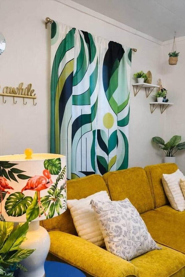 Une pièce avec un canapé jaune, une lampe à motifs, un rideau abstrait vert et blanc, et des étagères murales ornées de plantes.