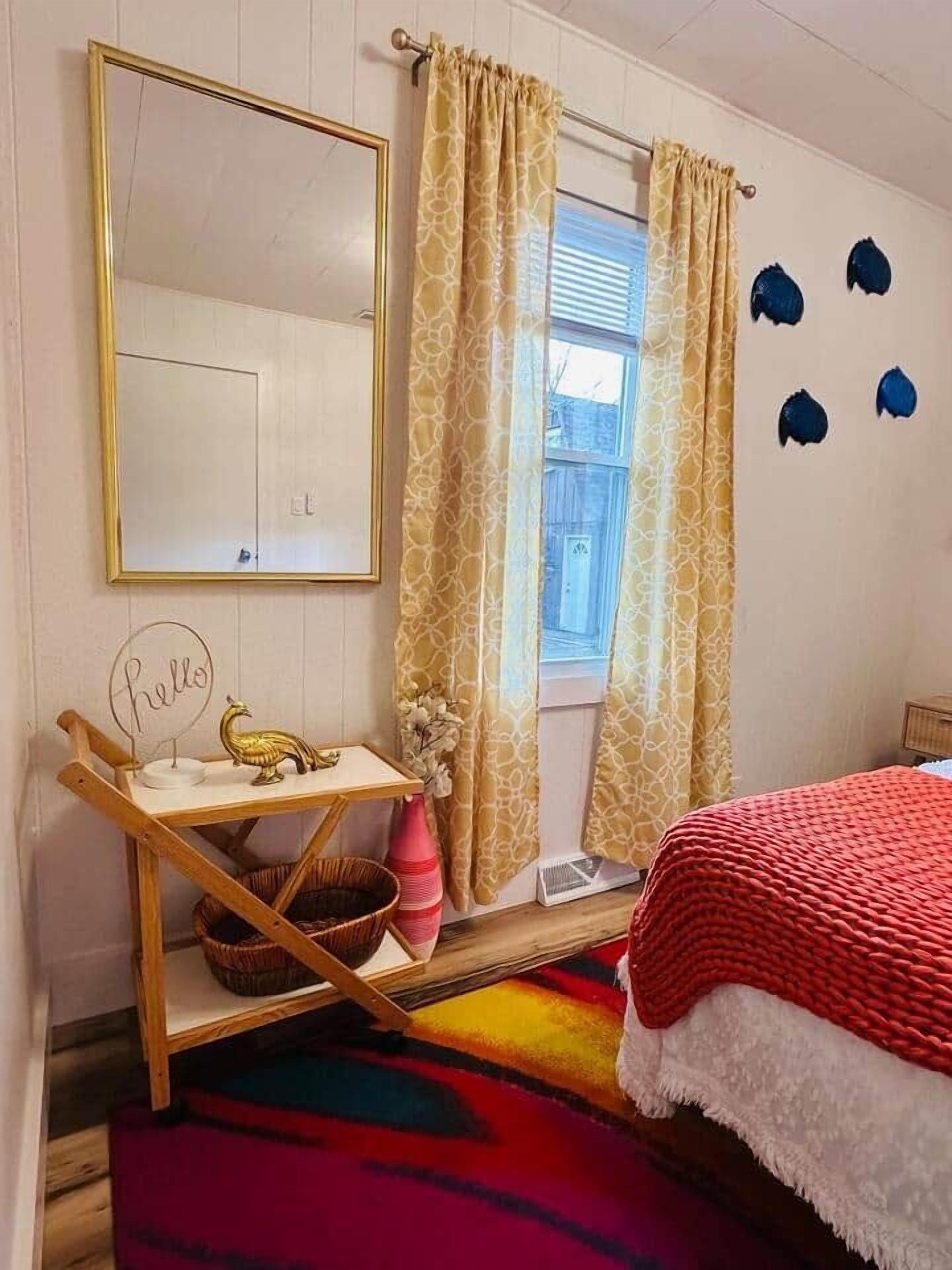 A bedroom with a bed, a window with curtains, a mirror, a bedside table with a decorative item, and a colorful rug.