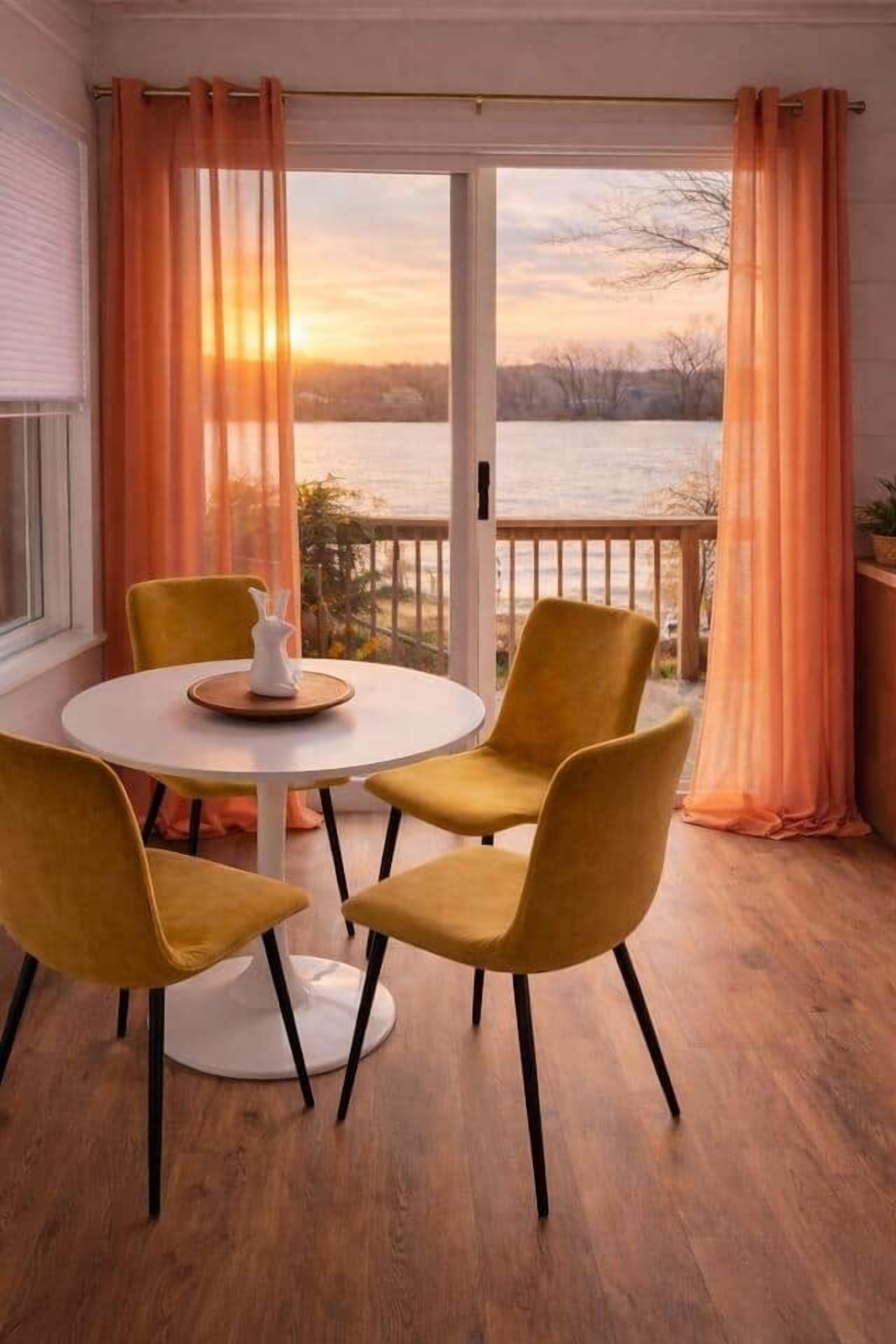 Comedor con una mesa redonda y cuatro sillas, una jarrón blanco y vista a un cuerpo de agua a través de una ventana con cortinas naranjas.