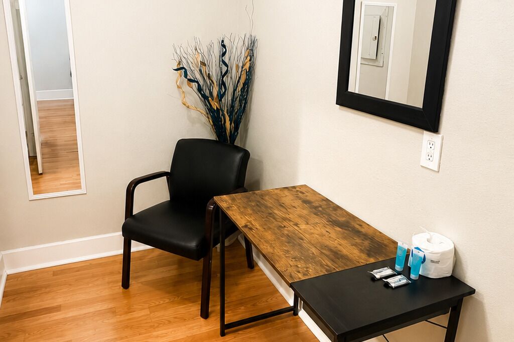 Una habitación con suelo de madera, una silla negra, una mesa de madera, un espejo y un jarrón con ramas decorativas.