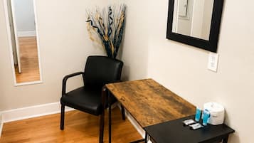 Una habitación con suelo de madera, una silla negra, una mesa de madera, un espejo y un jarrón con ramas decorativas.