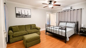 Une chambre à coucher avec un lit, un canapé vert, un pouf assorti, un ventilateur de plafond et une fenêtre avec des rideaux.