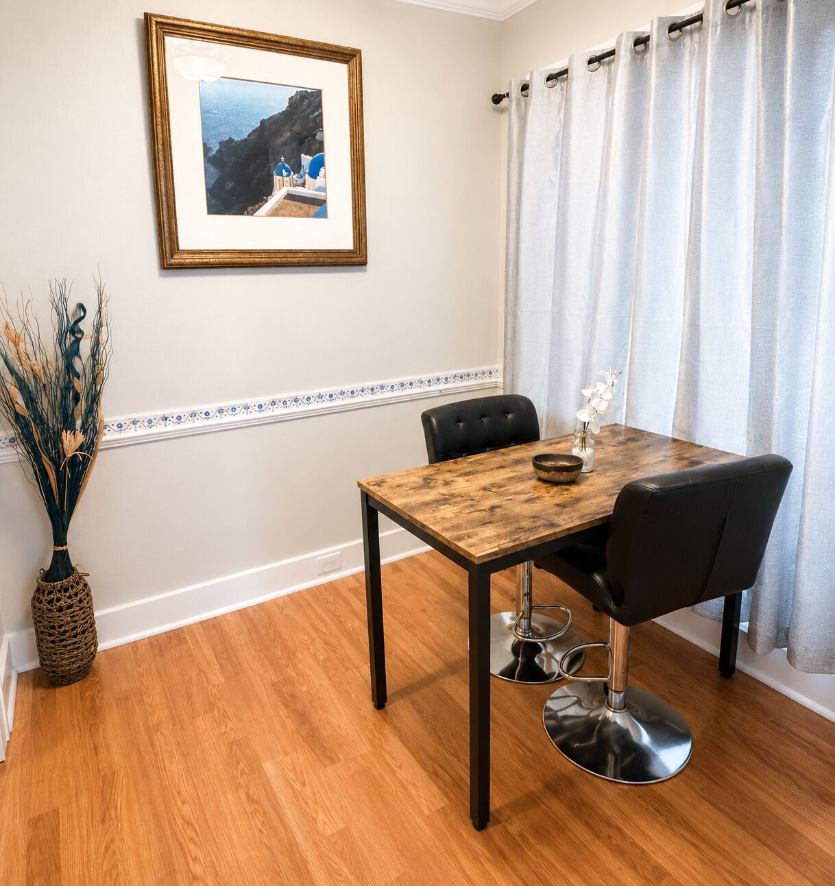 A dining area with a wooden table, black chairs, a framed picture on the wall, and a vase with dried plants.