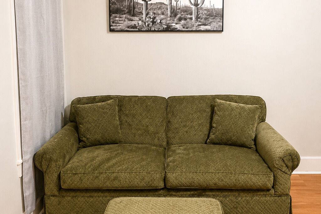 A green sofa with a matching ottoman in a room with a plain wall and a framed picture above.