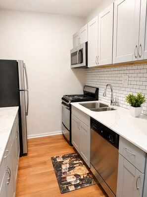 A compact kitchen with stainless steel appliances, white cabinets, a tiled backsplash, and a wooden floor. - 1BR Apartment - Walk to Toyota Center (Houston)