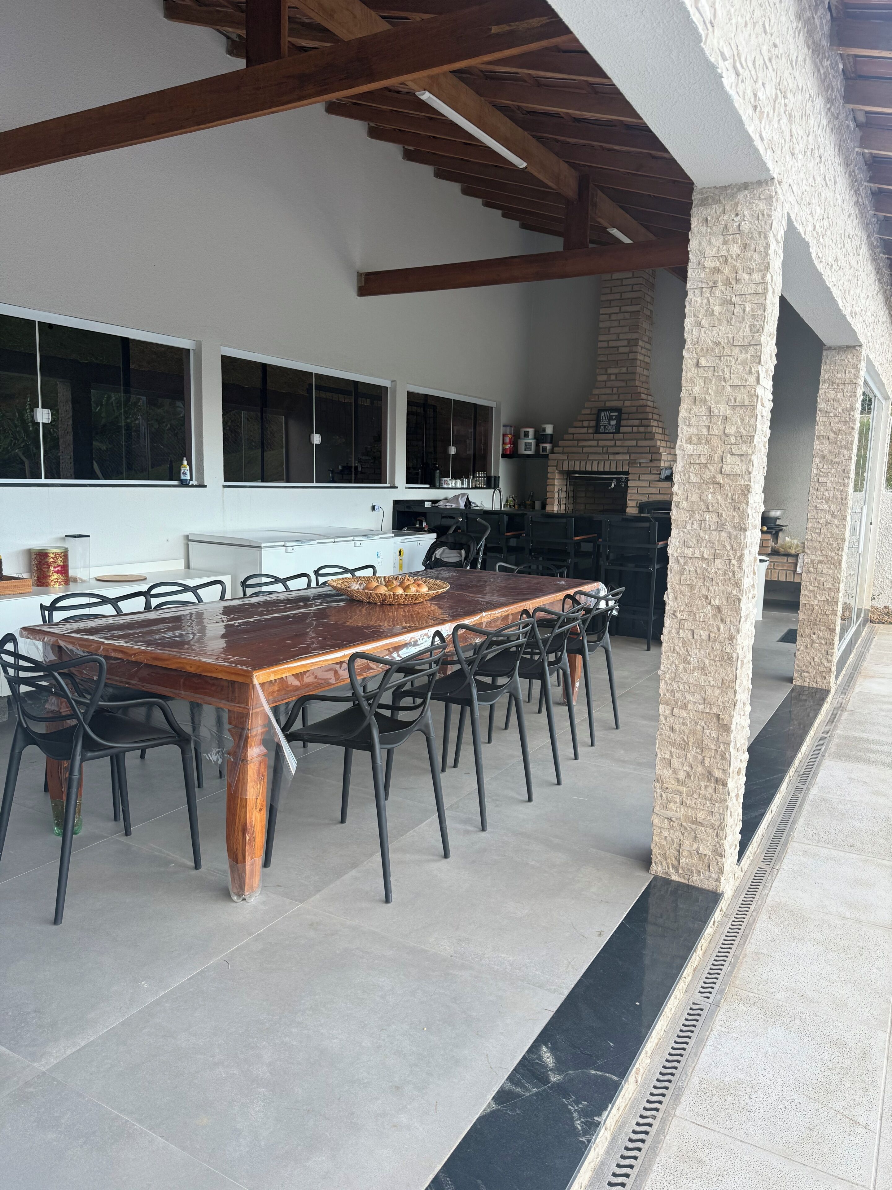 Un espace de restauration en plein air avec une grande table et des chaises en bois, un pilier en pierre et une cheminée en briques.