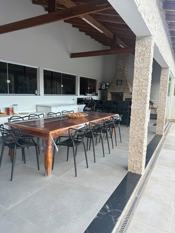 An outdoor dining area with a large wooden table and chairs, a stone pillar, and a brick fireplace.