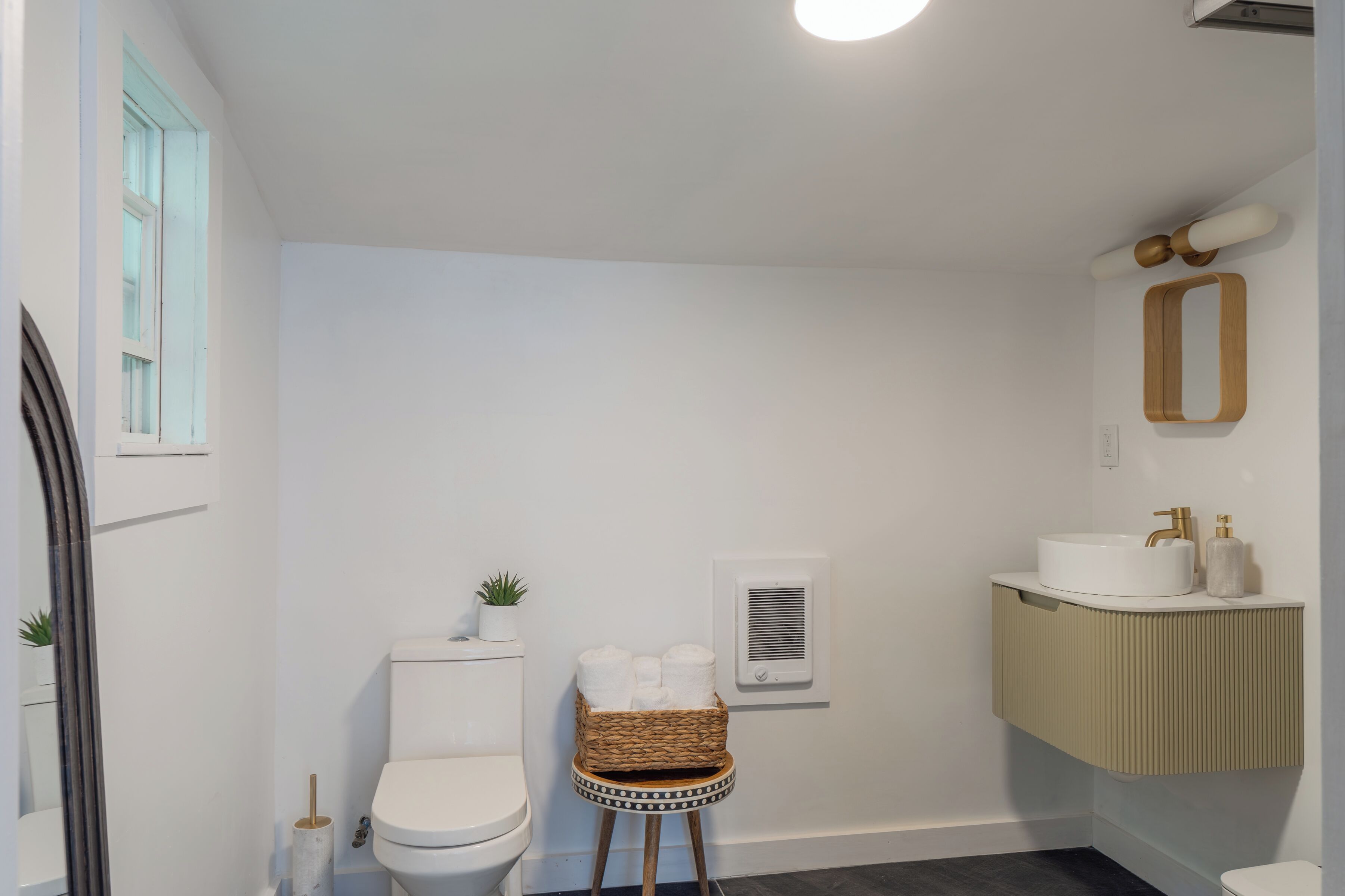 Ein Badezimmer mit Toilette, einem kleinen Korbwäscheschrank mit Handtüchern, einem wandmontierten Spiegel und einem Waschbecken mit Goldarmatur.