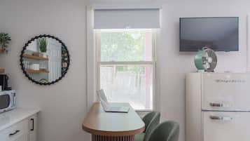 A room with a laptop on a wooden table, a television on the wall, a refrigerator, and a window with a view of greenery.