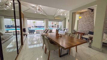 Ampla sala de estar com uma grande mesa de jantar de madeira, assentos confortáveis e vista para uma piscina ao ar livre.