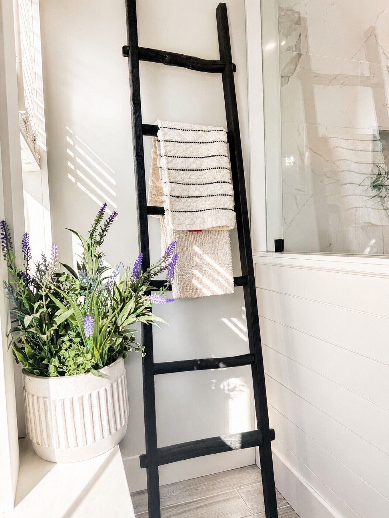 Une salle de bain avec une échelle noire, une serviette blanche et une plante en pot aux fleurs violettes.