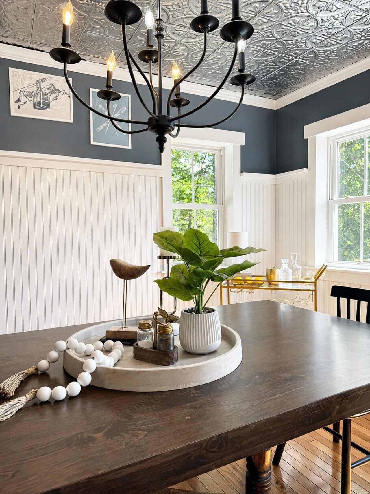 A dining area with a dark wooden table, white paneled walls, a black chandelier, and a large window.