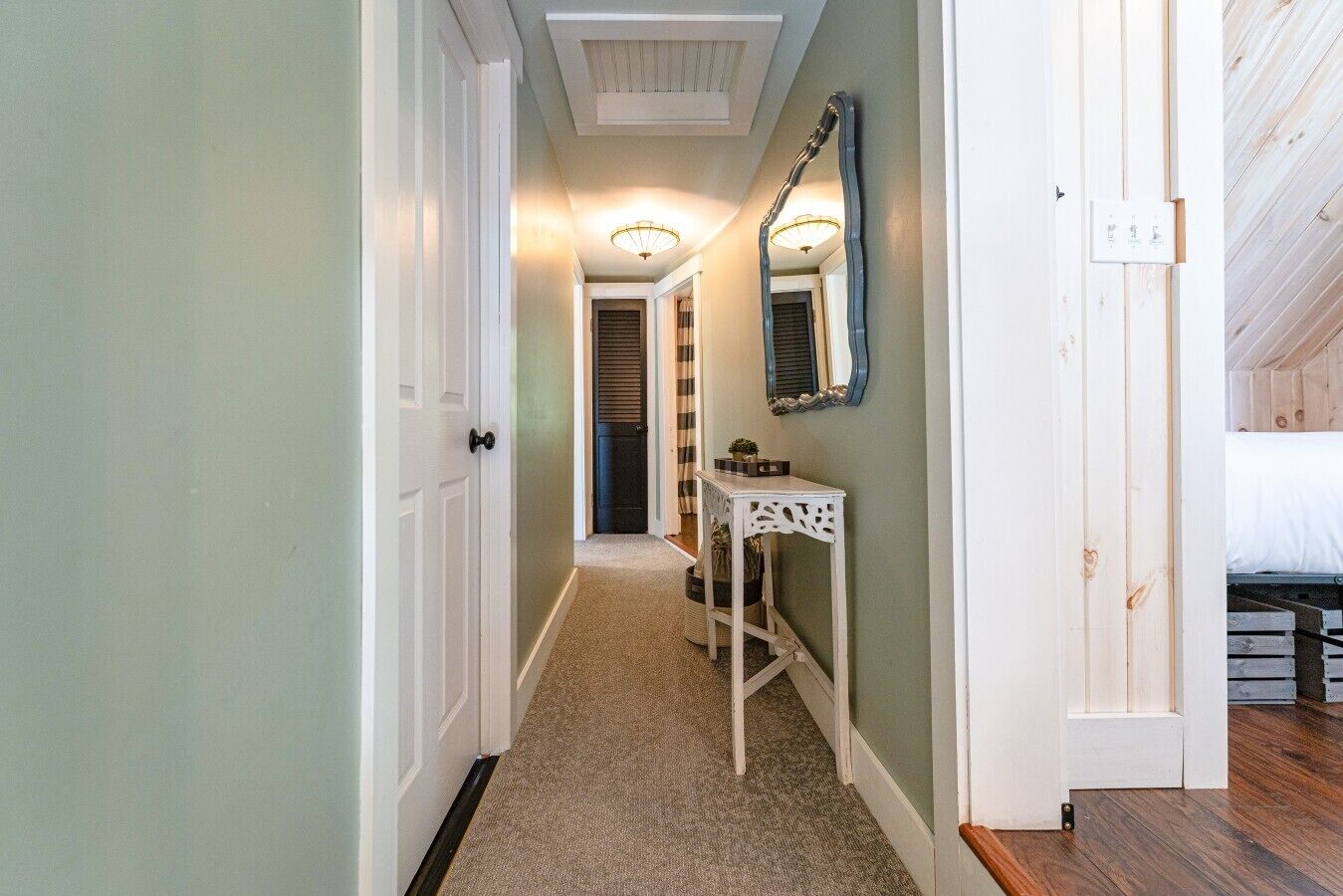 A hallway with a carpeted floor, a white door, a small table with a mirror, and a bed in the adjacent room.