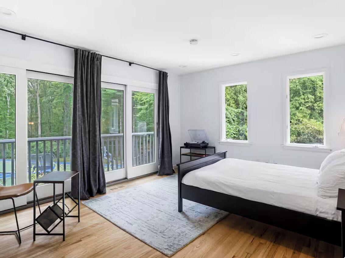 A bedroom with a bed, a desk, a chair, and large windows with curtains.