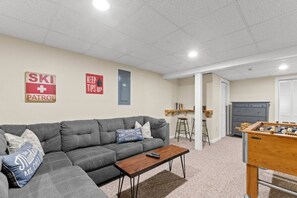 Ein Wohnzimmer mit einer Couch, einem Couchtisch und einem Kicker. Zwei Schilder an der Wand, eines mit der Aufschrift 'SKI + PATROL', das andere mit 'Tipps bitte hier lassen'.