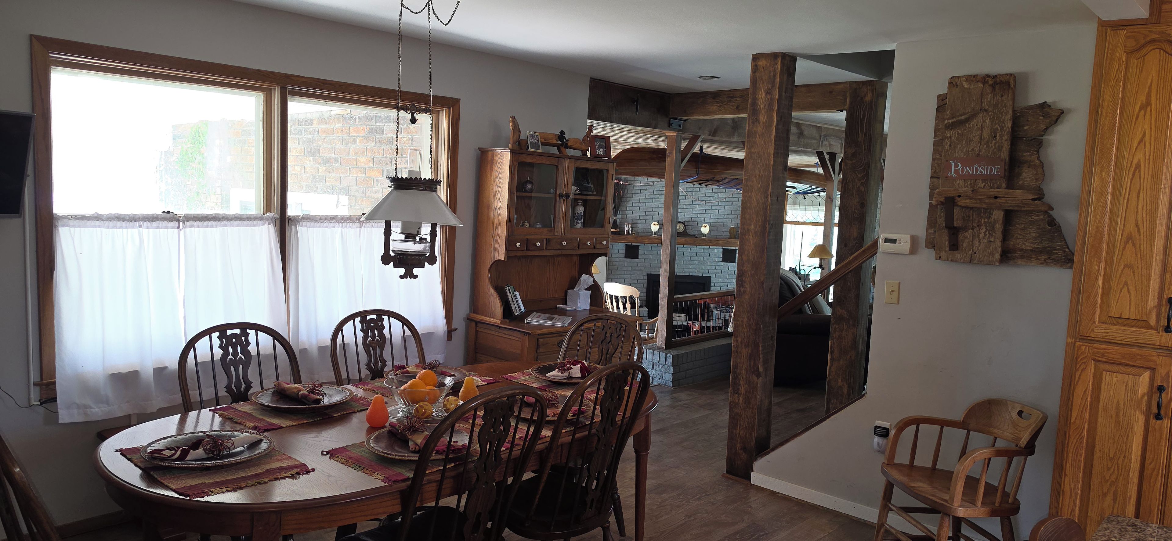 A dining area with a wooden table set for four, surrounded by chairs. There is a window with sheer curtains, a hanging lamp, and a brick wall in the background.