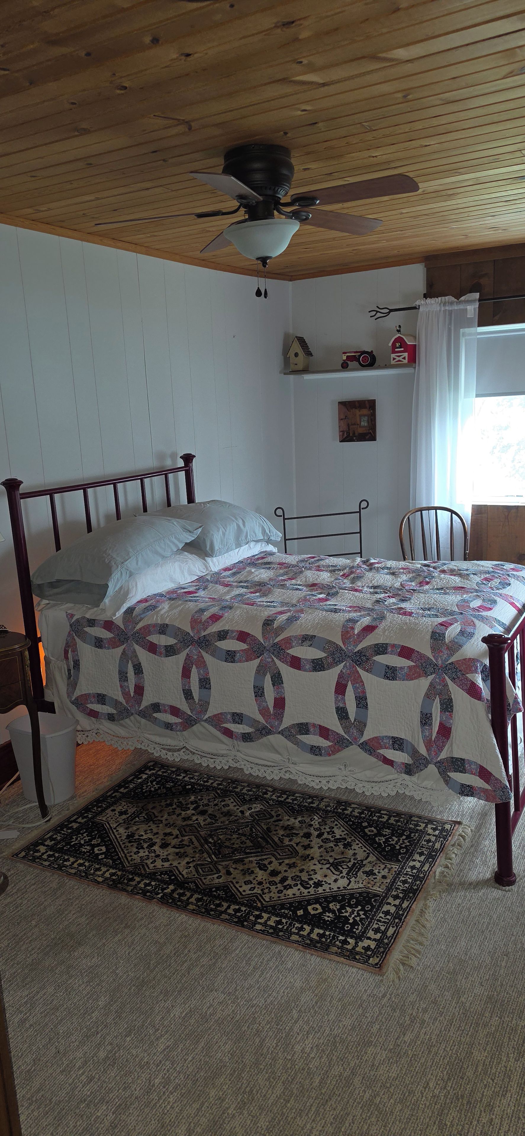 A bedroom with a bed, a ceiling fan, a window with curtains, and a small shelf with decorative items.