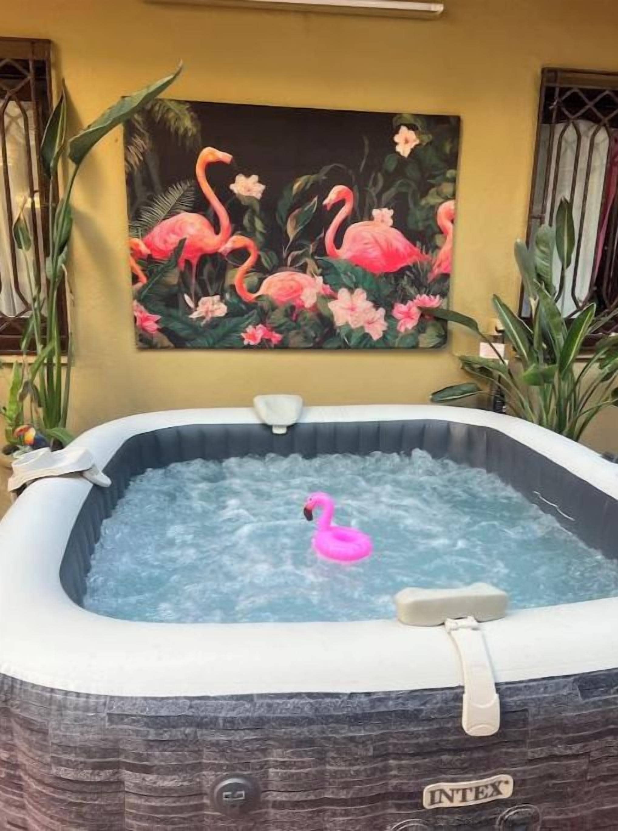 Un bain à remous avec un flotteur en forme de flamant rose, entouré de plantes, et une peinture représentant des flamants roses sur le mur.