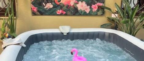 Un bain à remous avec un flotteur en forme de flamant rose, entouré de plantes, et une peinture représentant des flamants roses sur le mur.