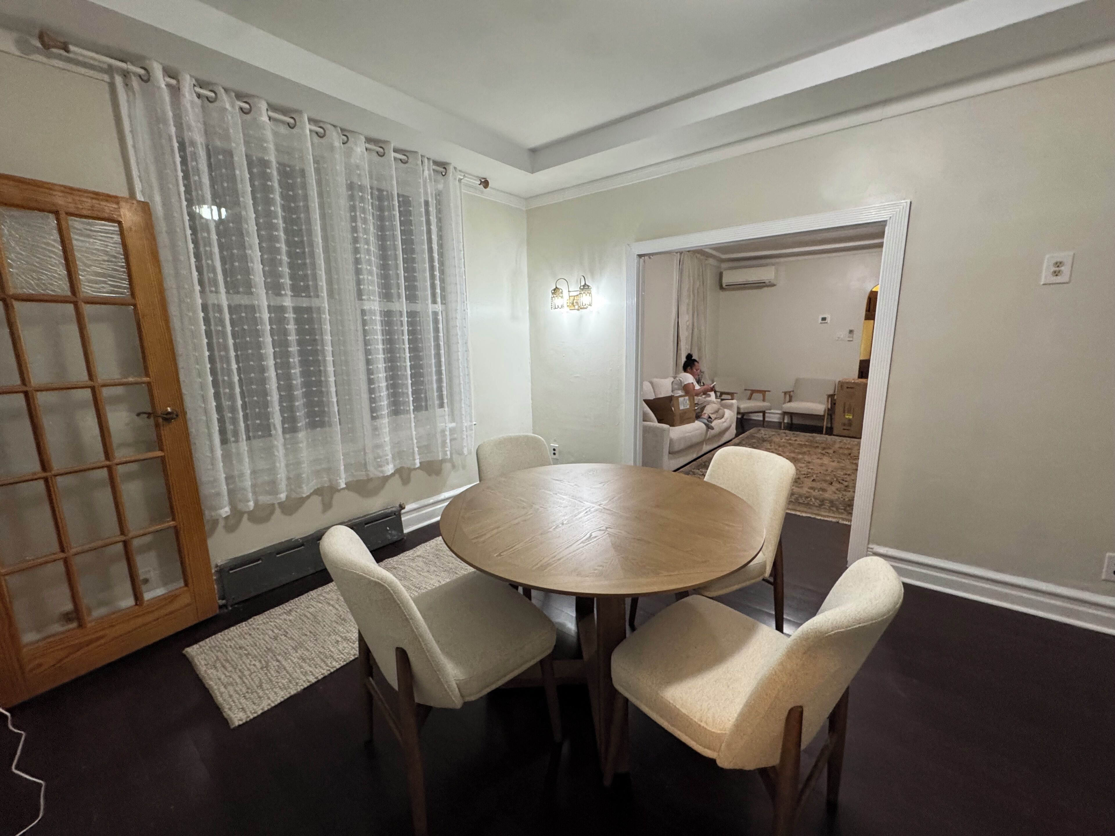A dining area with a round table and four chairs, a wooden door with glass panels, and a living room in the background.