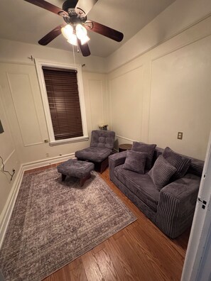 Ein Wohnzimmer mit Deckenventilator, grauem Sofa, Hocker und einem gemusterten Teppich.