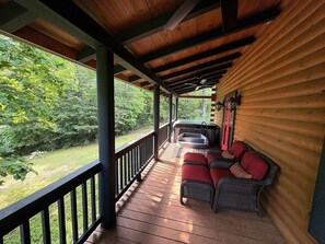 Eine überdachte Veranda mit Holzterrasse, eine Sitzecke mit Korbmöbeln und roten Kissen sowie ein Blick auf eine grüne Landschaft.