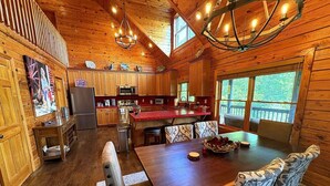 A wooden cabin interior with a dining area, kitchen, and large windows. - Authentic Log Cabin • Hot Tub, Game Room & Yard Games • Sleeps 12 • Wine Country (Mount Airy)