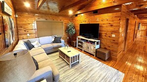 A cozy log cabin interior with a sectional sofa, a flat-screen TV, and a wooden ceiling. - Authentic Log Cabin • Hot Tub, Game Room & Yard Games • Sleeps 12 • Wine Country (Mount Airy)