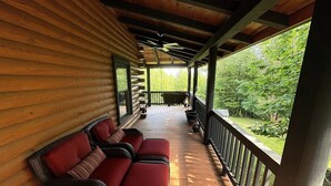 A covered porch with wicker furniture and a view of the surrounding greenery. - Authentic Log Cabin • Hot Tub, Game Room & Yard Games • Sleeps 12 • Wine Country (Mount Airy)