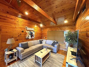 A cozy cabin interior with a sectional sofa, a small table, a lamp, and a television.