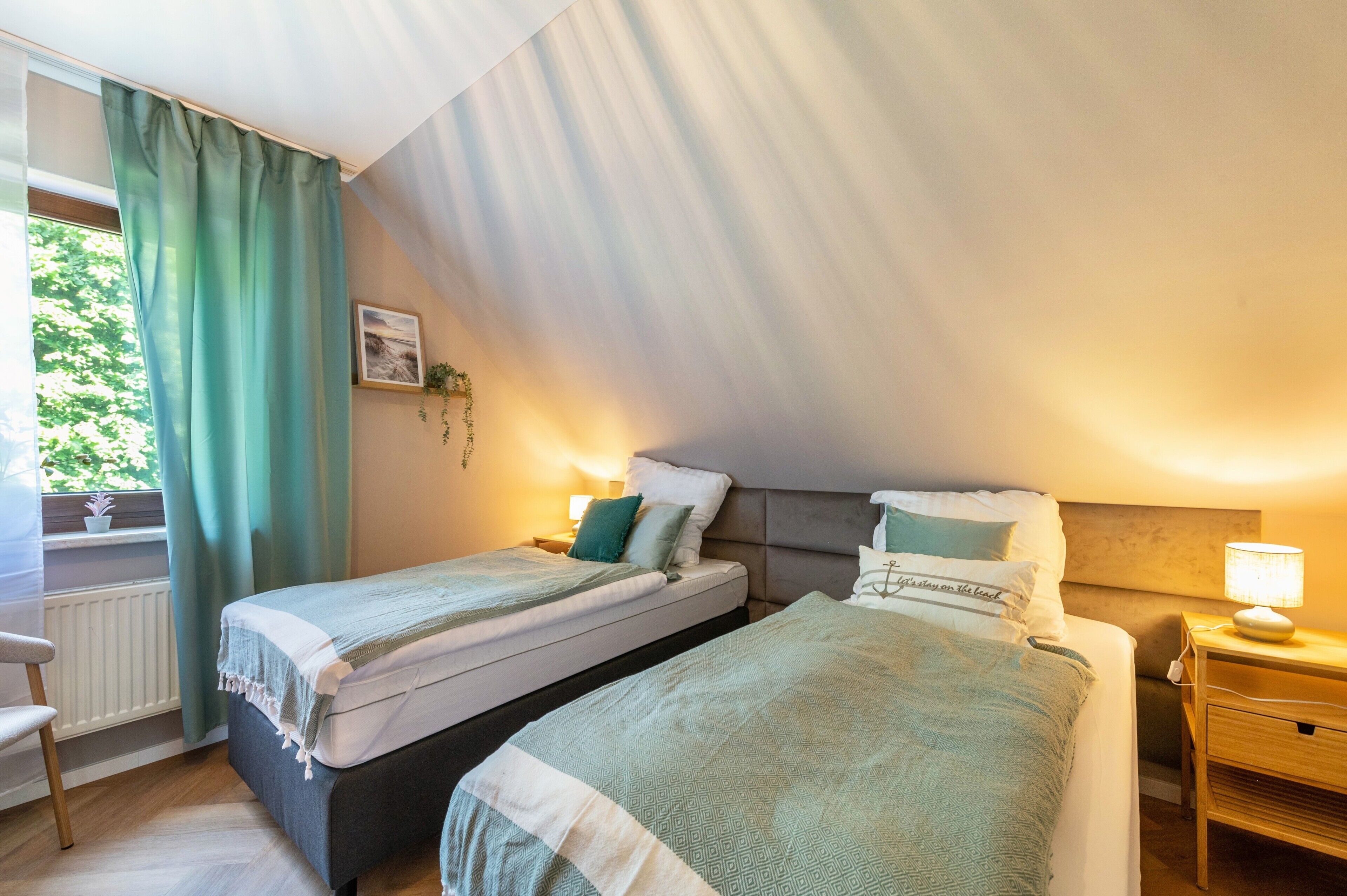 A bedroom with two beds, a window with curtains, a bedside table with a lamp, and a radiator.