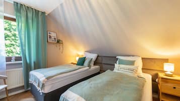 A bedroom with two beds, a window with curtains, a bedside table with a lamp, and a radiator.