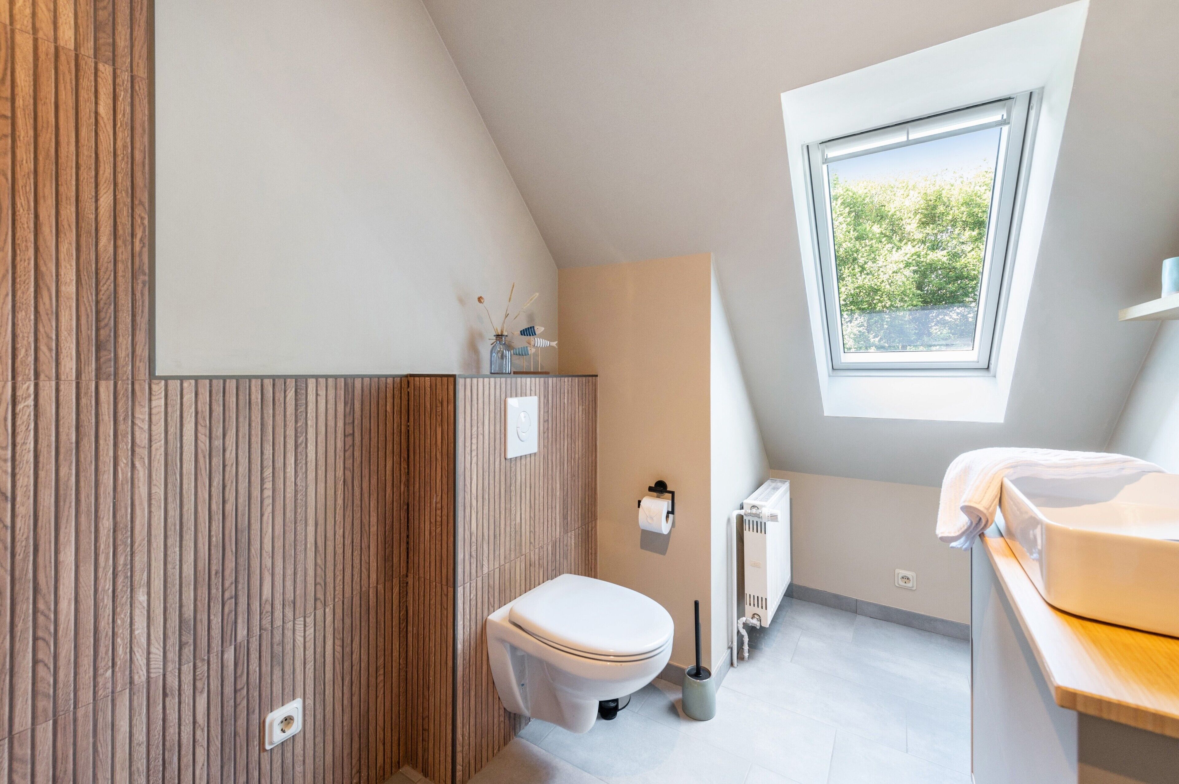A modern bathroom with a wooden panel wall, a toilet, a radiator, and a window providing natural light.