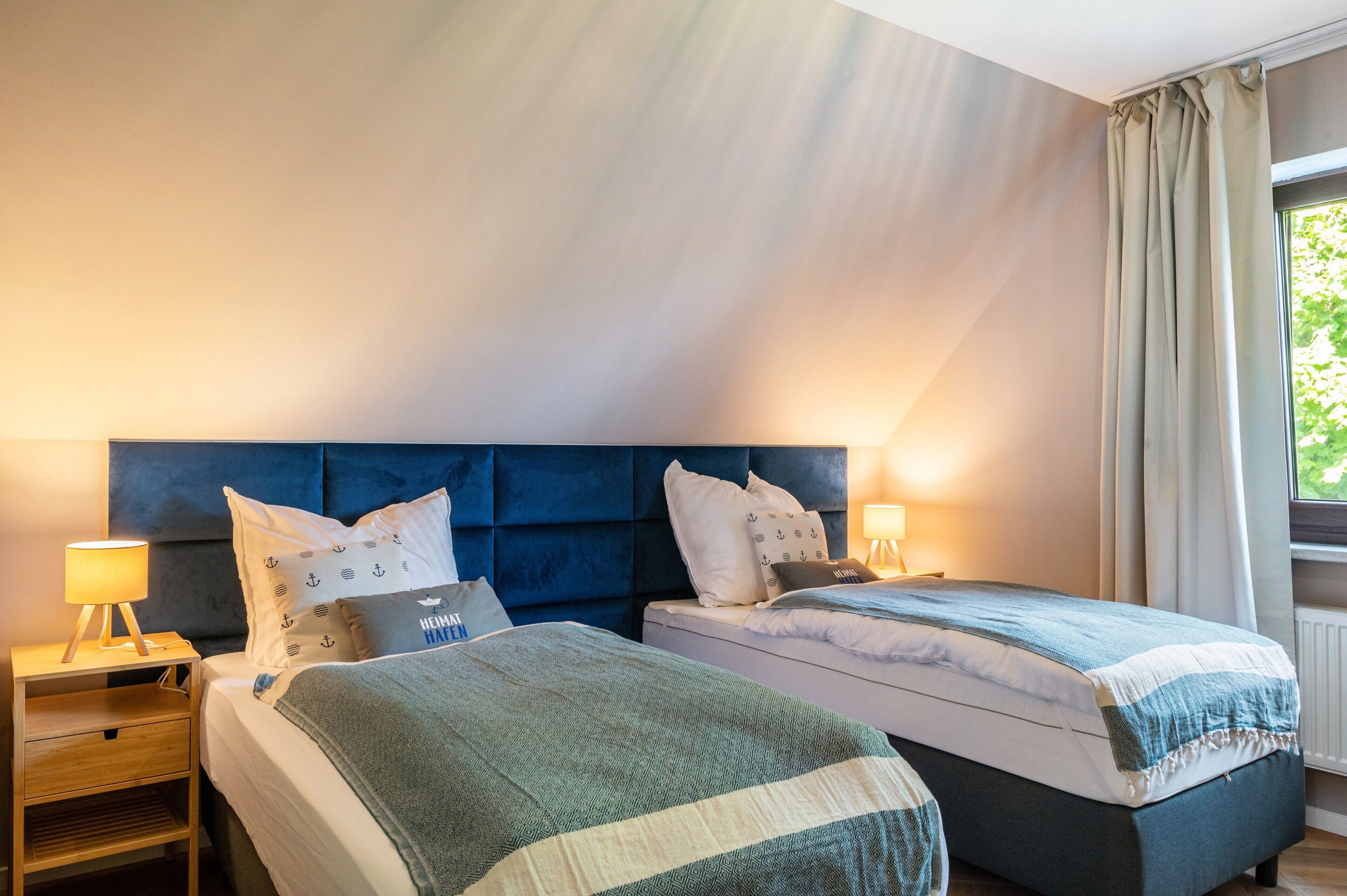 A hotel room with two beds, a nightstand with a lamp, and a window with curtains.