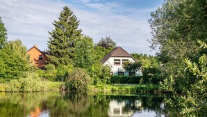 A tranquil riverside scene with two houses, one with a brown roof and another with a white facade, surrounded by lush greenery and a calm water body reflecting the structures.