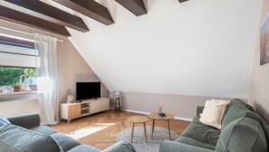 A modern living room with a sloped ceiling, a flat-screen TV, a wooden cabinet, a small round table, and a green sofa. - Apartment "Kleiner Anker" with Private Terrace, Garden, and Wi-Fi (Papendorf)