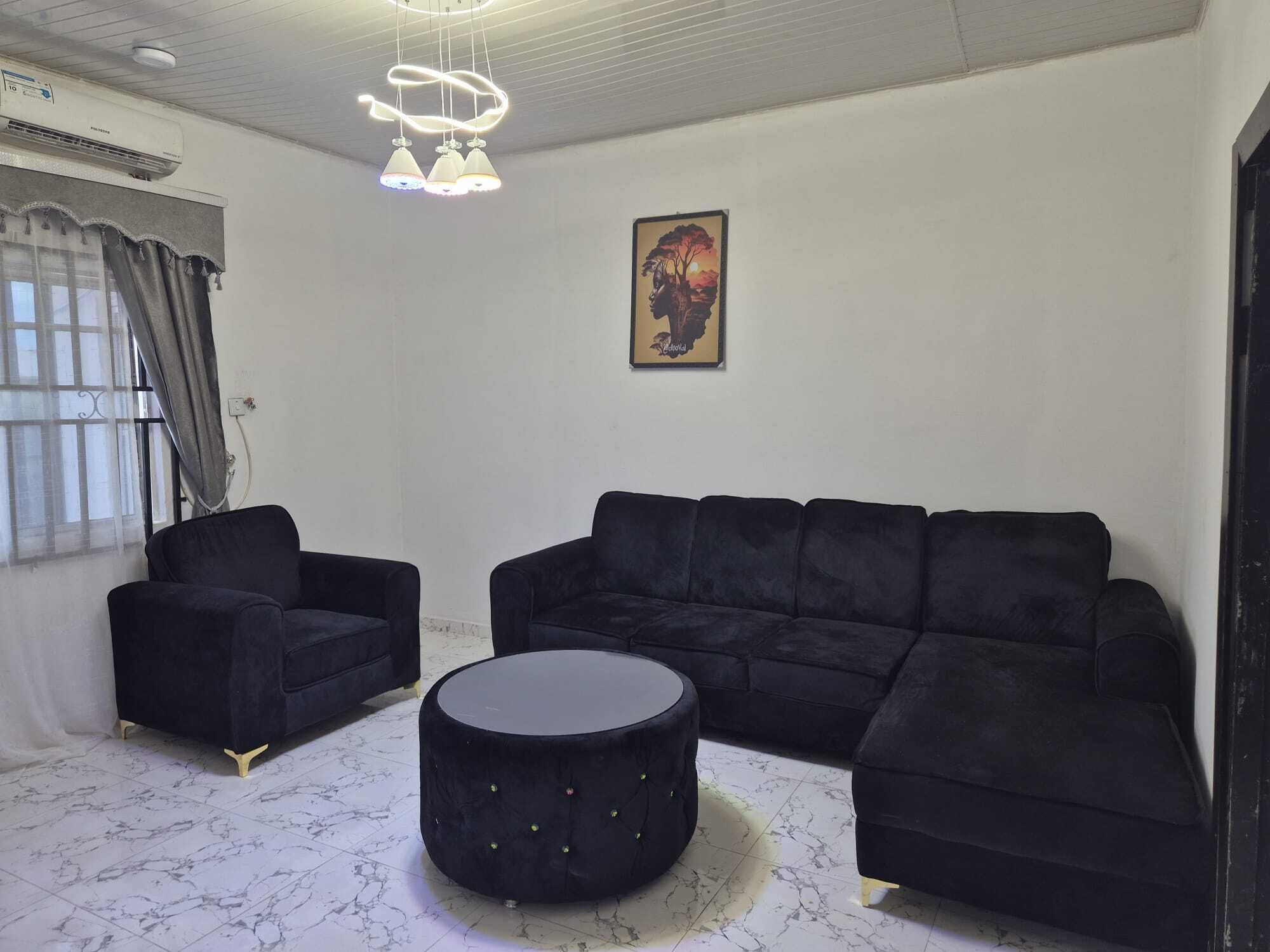 A modern living room with a black sofa set, a round ottoman, and a pendant light fixture.