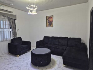 Ein modernes Wohnzimmer mit einer schwarzen Sofagarnitur, einem runden Hocker und einer Pendelleuchte.