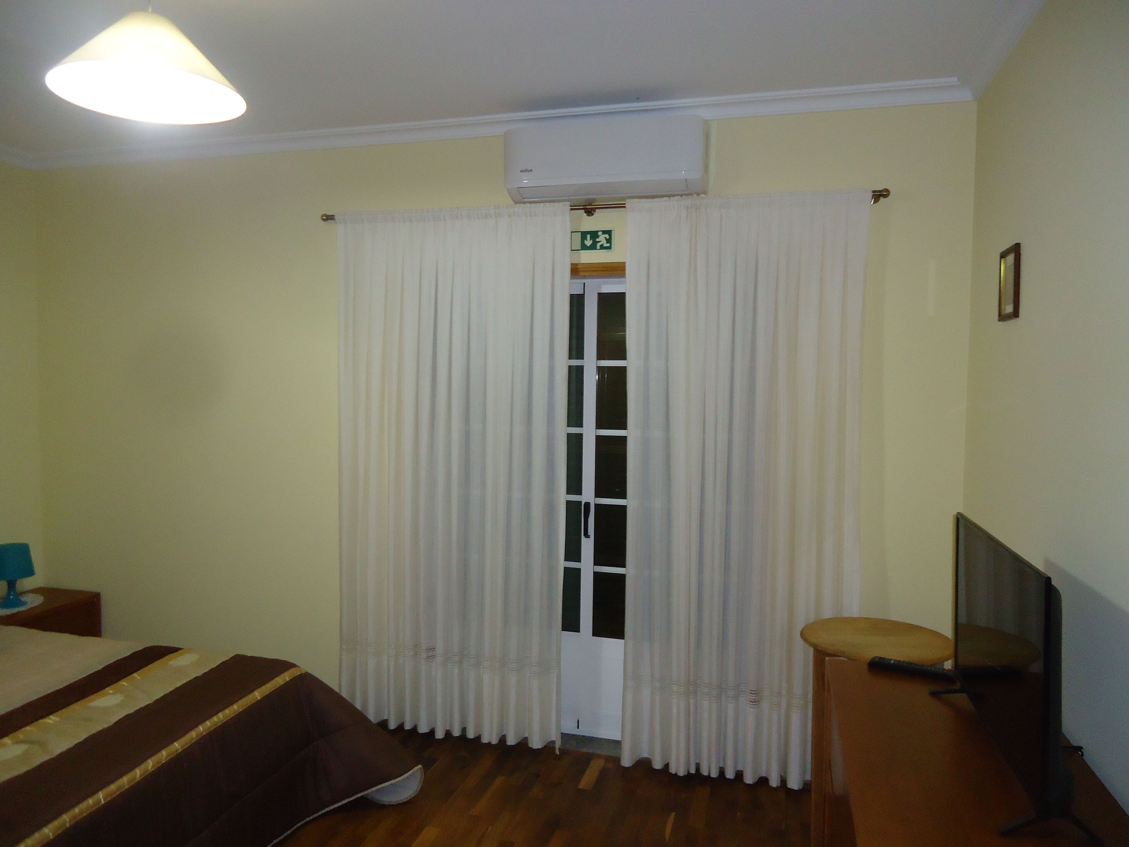 Une chambre d’hôtel équipée d’un lit, d’une télévision, d’une chaise, d’une fenêtre avec des rideaux, d’un climatiseur et d’une porte portant un panneau indiquant la sortie de secours.