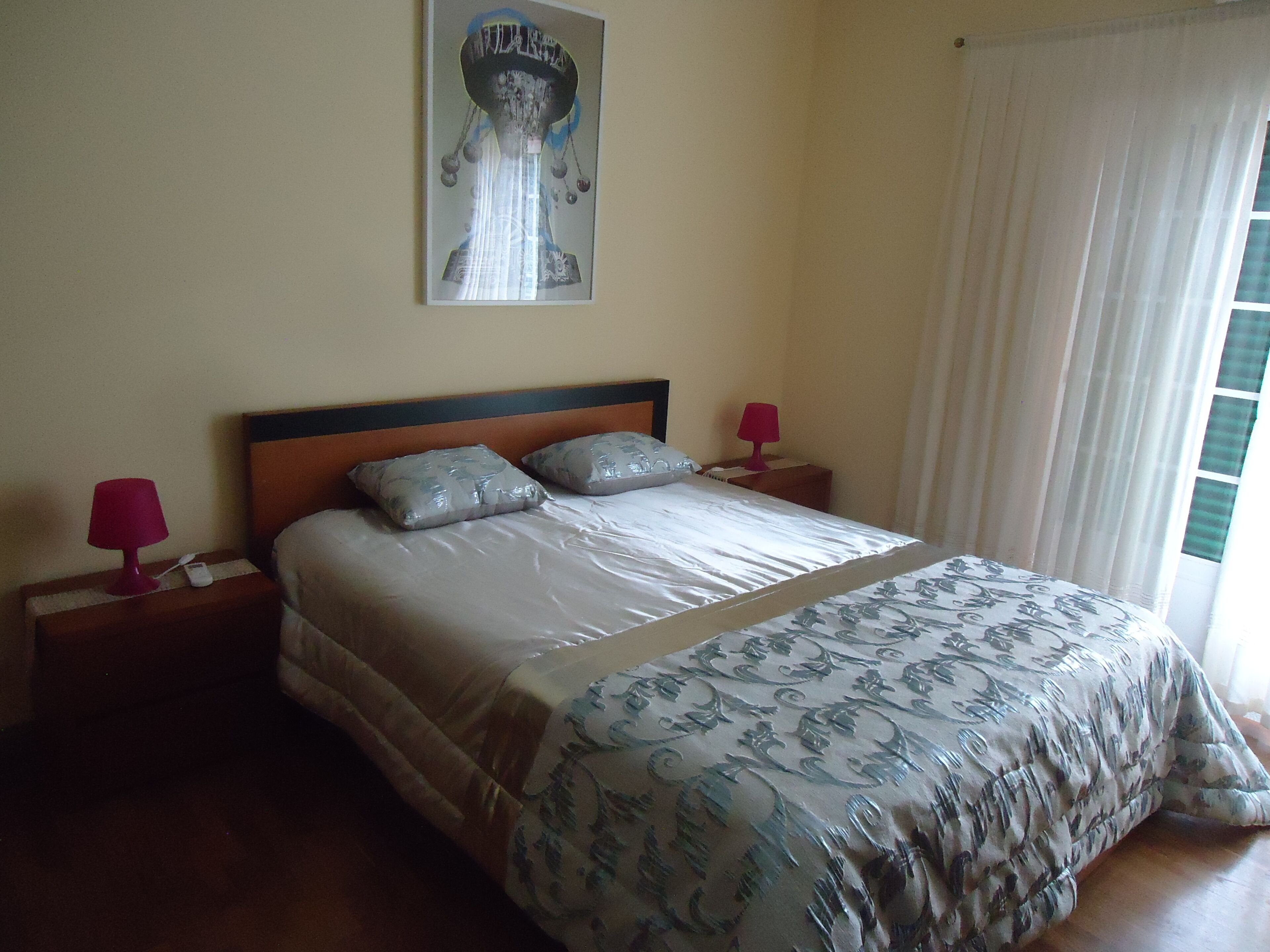 Una cama bien hecha con una colcha estampada, dos almohadas y un cabecero. Se ven dos mesitas de noche con lámparas y un cuadro colgado en la pared.