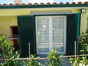A building with a green door and shutters, a small brick structure to the left, and a tiled roof.