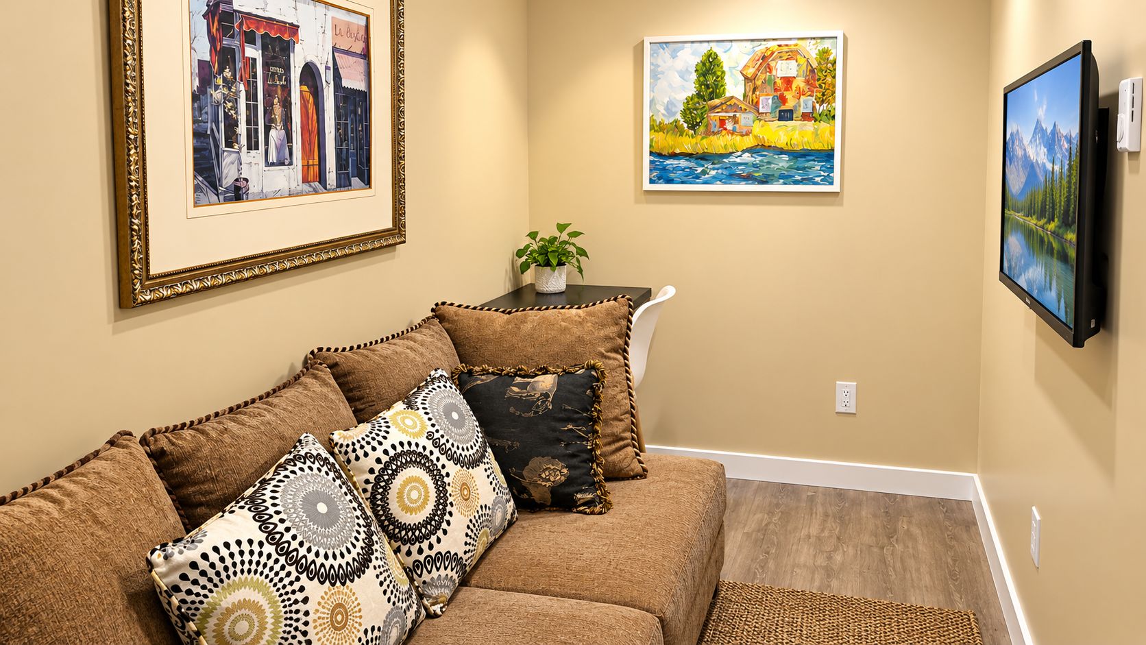 A living room with a brown sofa, patterned cushions, a small table with a plant, and two framed artworks on the walls.