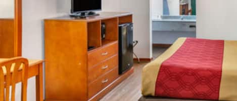 A hotel room with a wooden dresser, a television, a bed with a red and beige bedspread, and a chair.