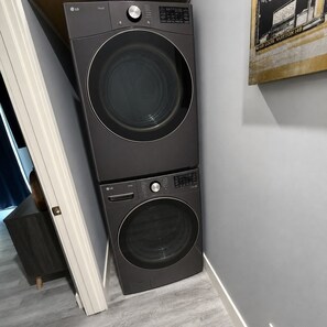 En svart tvättmaskin av märket LG THING står i ett hörn av ett rum med en vit dörr och ett mörkt träskåp.