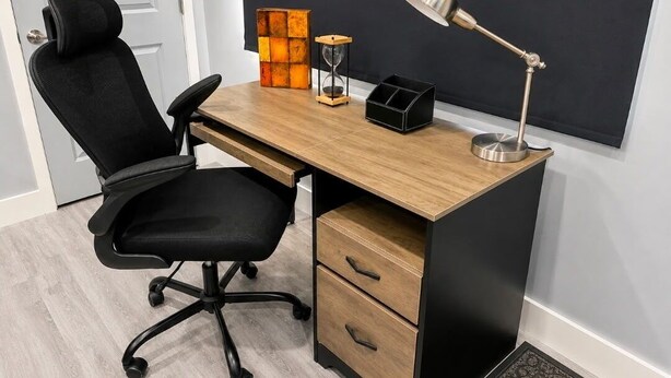 Ein moderner Schreibtisch mit schwarzem Bürostuhl, einer Holzschreibtischplatte, einer schwarzen Aufbewahrungsbox, einer metallischen Schreibtischlampe und einer dekorativen Sanduhr.
