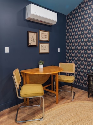 A dining area with a round wooden table, two chairs, a potted plant, and framed botanical prints on the wall.
