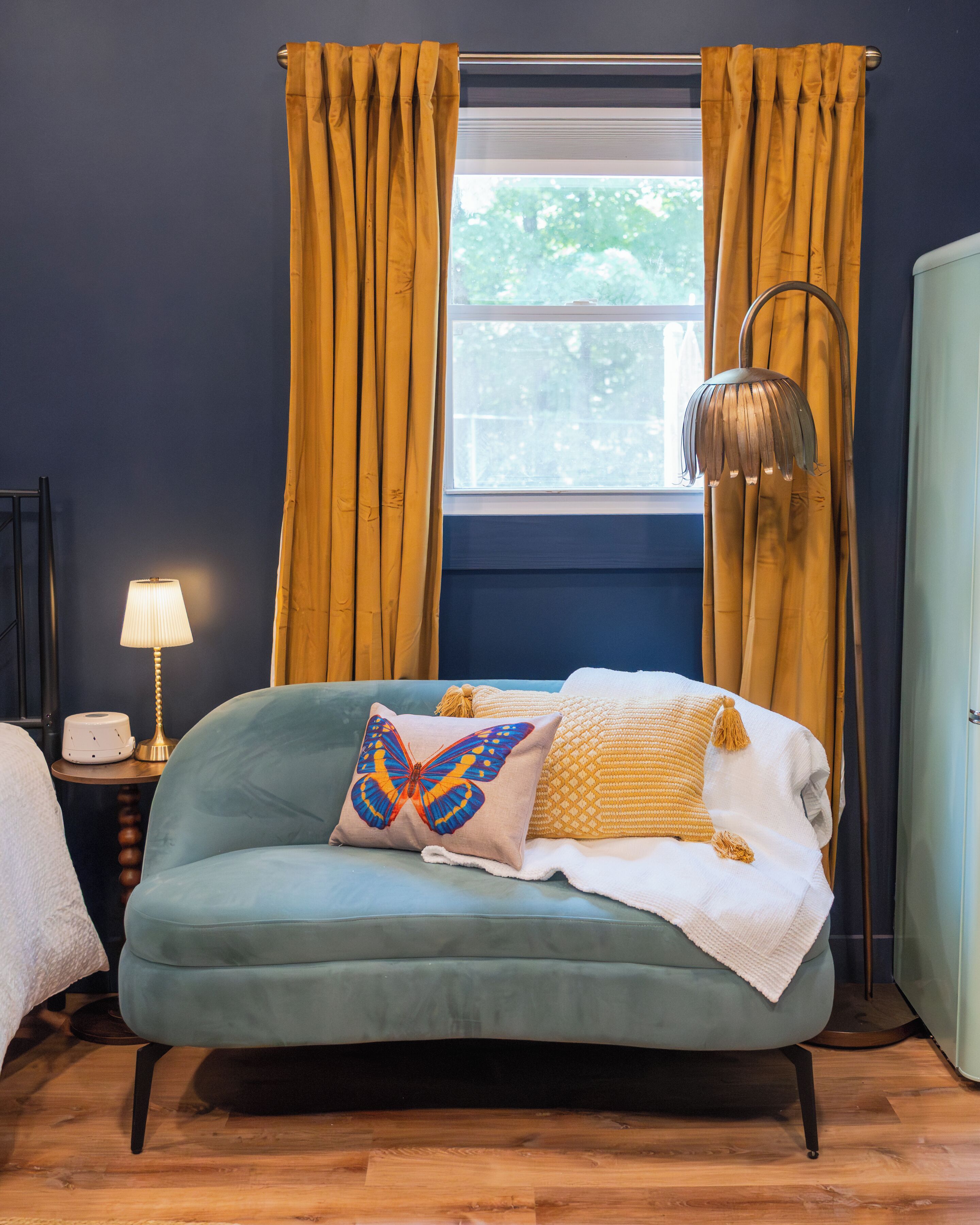 Une pièce avec un canapé de couleur turquoise, une fenêtre ornée de rideaux jaunes, un lampadaire et un lit recouvert d’une couverture blanche.